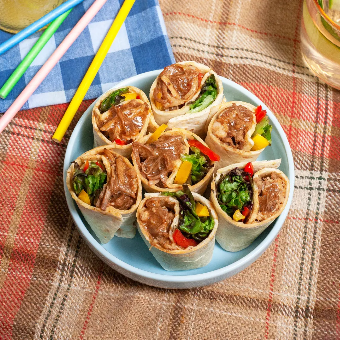 A blue bowl atop a picnic blanket. The bowl is filled with pulled pork tortilla wraps.